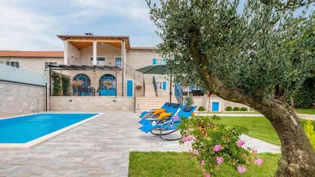 Un espace piscine avec des chaises longues, une terrasse en pierre et un bâtiment aux portes et colonnes bleues.