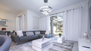 Ein modernes Wohnzimmer mit grauem Sofa, weißem Couchtisch und einem Glastisch. Eine Pendelleuchte, eine Pflanze und ein Spiegel sind an der Wand zu sehen.