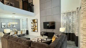 Een ruime woonkamer met een grote bank, een eethoek met een kroonluchter, en een flatscreen televisie aan de muur.