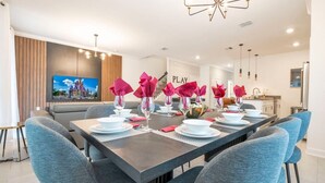 A modern dining area with a long table set for six, featuring grey upholstered chairs and a dark table. A large flat-screen TV is mounted on the wall, displaying a colorful image. - Stunning 4 Bd w Pool Storey Lake Resort 2400 (Kissimmee)