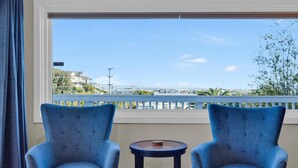 Ein Zimmer mit zwei blauen Polstersesseln und einem kleinen runden Tisch, ein großes Fenster mit Blick auf eine Stadtlandschaft und einem Balkongeländer.
