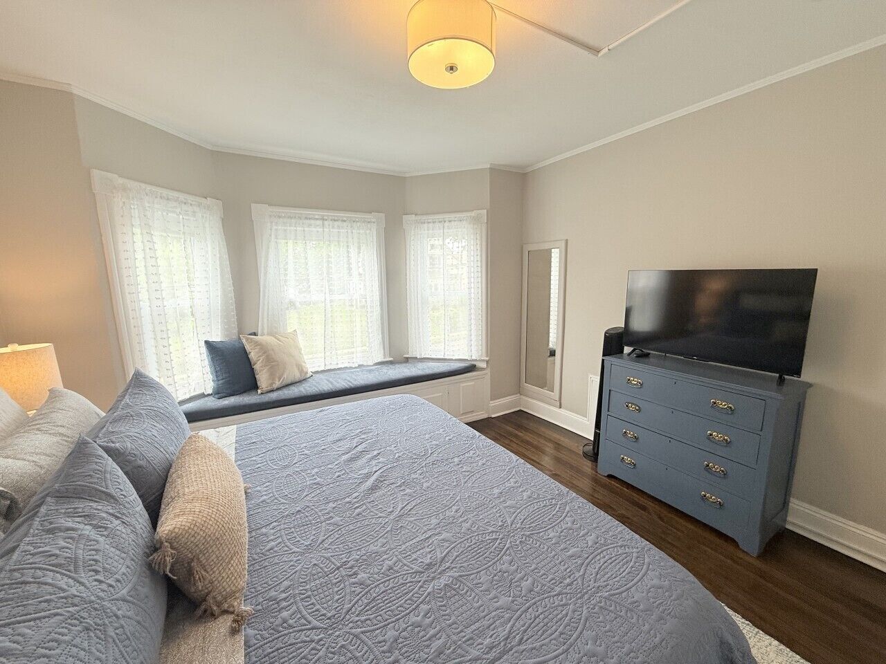 A bedroom with a bed, a bench, a dresser, a television, and a window with curtains.