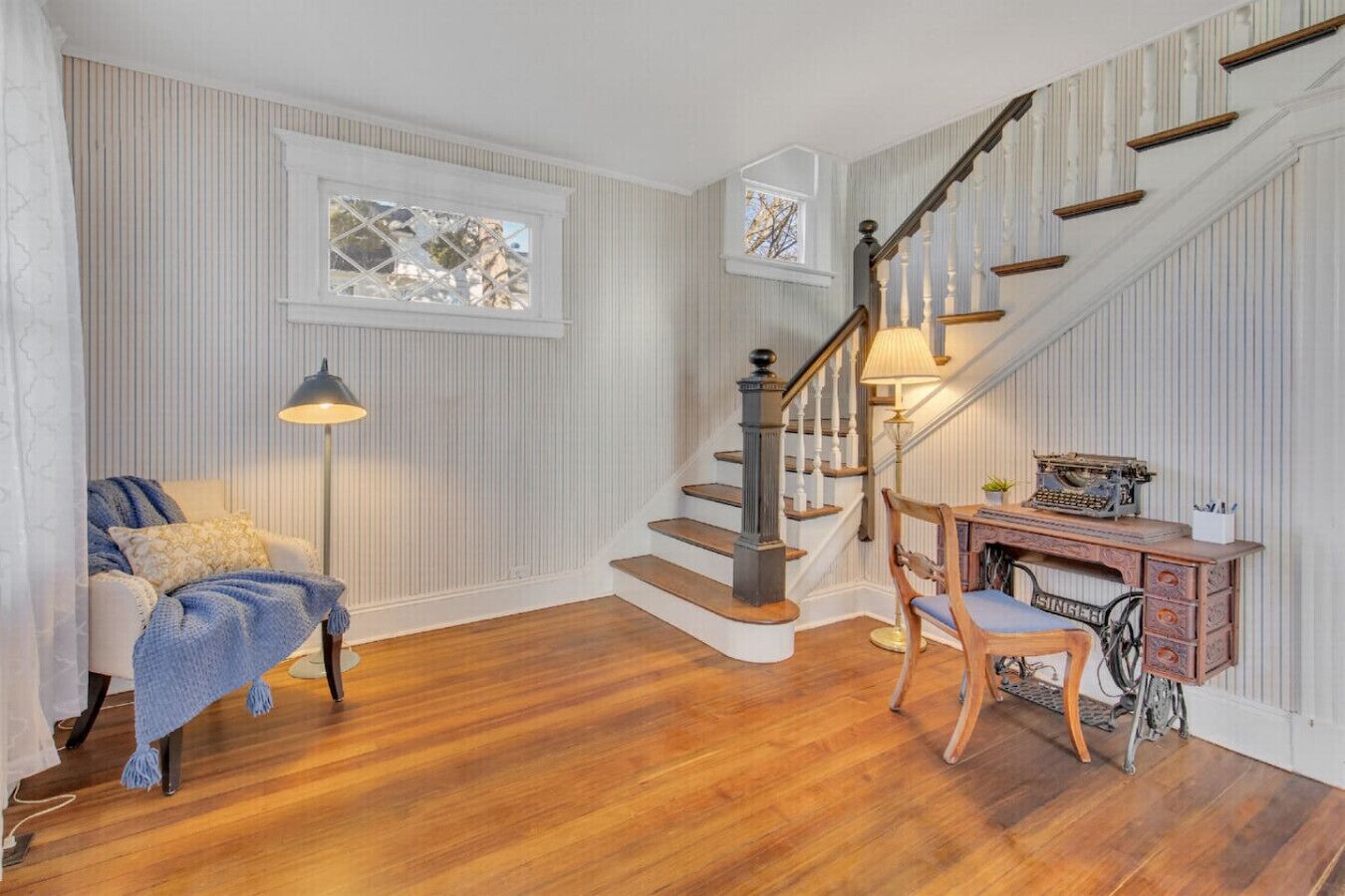 A room with a wooden staircase, a desk with a typewriter, a chair, a lamp, and a window with a view.