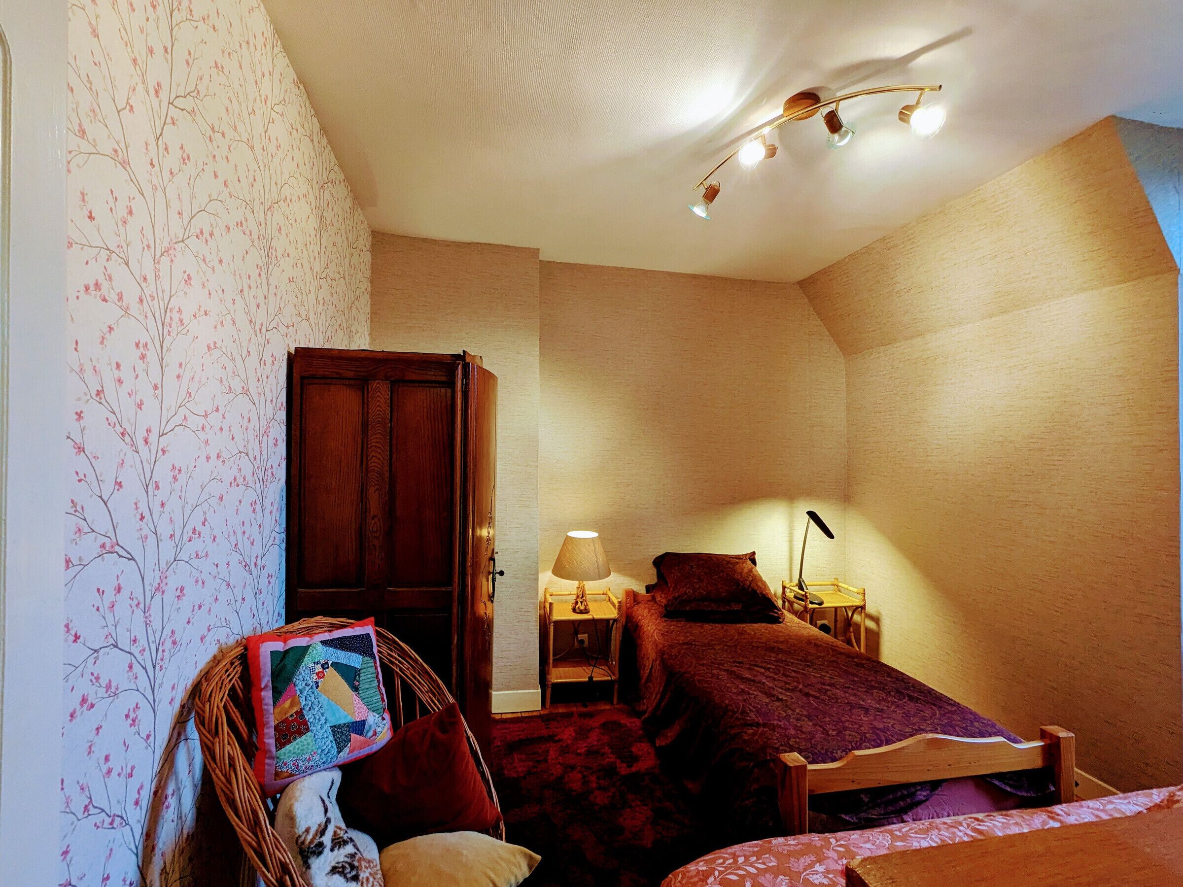 Ein Schlafzimmer mit einem Holzschrank, einem Bett mit roter Decke, einem Nachttisch mit Lampe und einer Wand mit floralem Wandpapier.