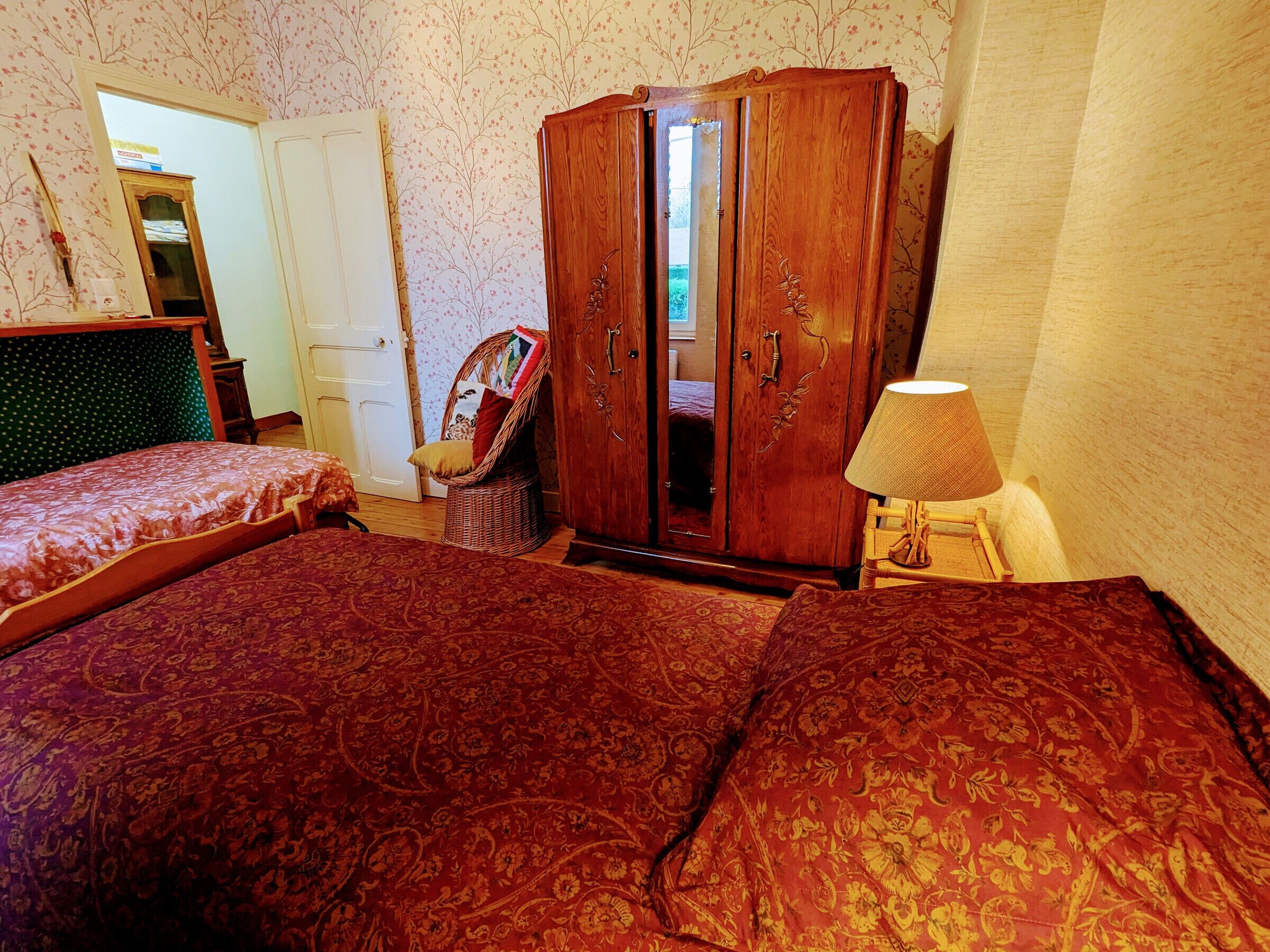 A bedroom with two beds, a wooden wardrobe, a wicker chair, and a bedside lamp.
