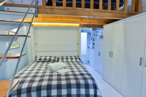 A bedroom with a checkered bedspread, a wooden ladder, a wardrobe, and a door leading to another room.