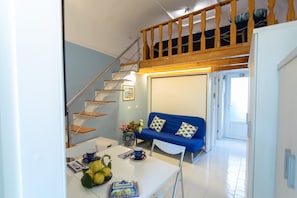 A compact living space with a staircase leading to a loft, a blue sofa with patterned cushions, and a dining area with a table set for two.