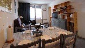 Un comedor con una mesa preparada para cuatro personas, un televisor sobre una estantería de madera y una ventana con cortinas.