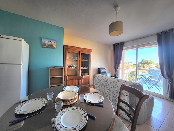 A dining area with a table set for four, a refrigerator, a cabinet with dishes, and a view of the outdoors.