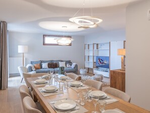Una sala de estar moderna con comedor listo para seis personas. Incluye un sofá, un televisor montado en la pared y una mesa de comedor de madera con sillas.