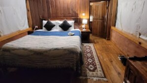 Une chambre avec un plafond en bois, un lit recouvert d’une courtepointe bleue, deux oreillers noirs, une table de chevet avec une lampe et une armoire en bois.