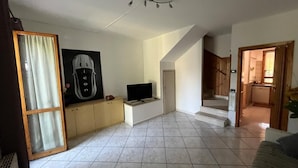 A spacious living area with a tiled floor, a flat-screen TV, a large framed picture, and a staircase leading to a kitchen.