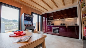 A modern kitchen with a view of the countryside, a wooden dining table, and a retro arcade machine. - Chalet Henri, Grand Confort, au Calme, vue D'exception, Sauna, 4/6 Personnes (Murol)
