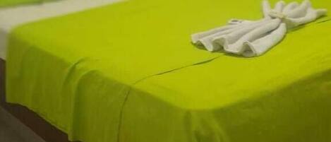 Una cama con una colcha verde y toallas blancas, cabecera a rayas y cortinas amarillas.