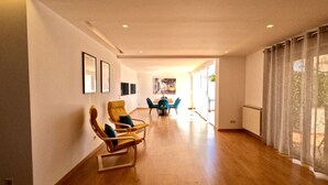 A spacious room with wooden flooring, a dining area with blue chairs, and a large window with sheer curtains. - Beloura (Sintra)