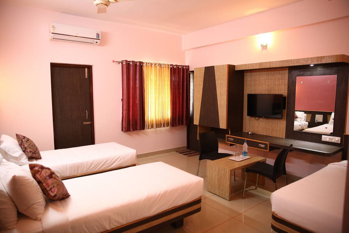 A hotel room with two single beds, a desk with a chair, a flat-screen TV mounted on the wall, and a small table with a bottle and glasses.