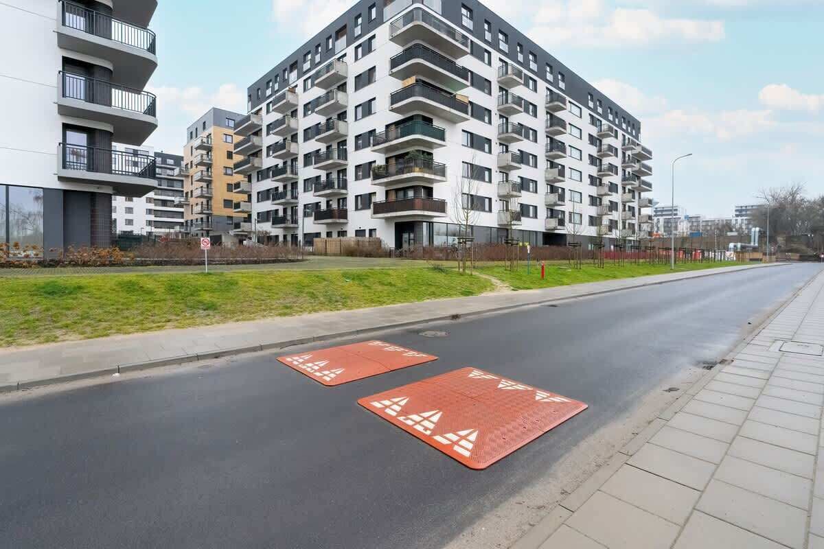 Un edificio residencial de varios pisos con balcones, una calle pavimentada con alfombras de seguridad rojas y una acera.