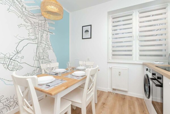 Un coin repas avec une table en bois, des chaises blanches, une carte accrochée au mur, un luminaire suspendu et une fenêtre avec des stores.