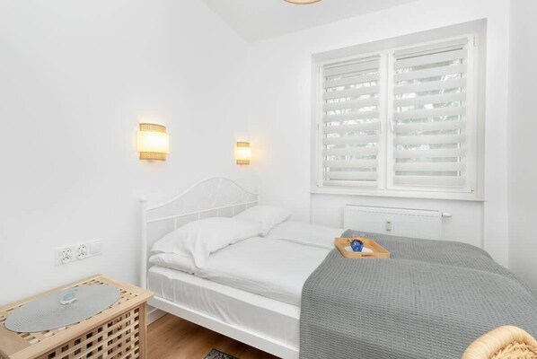 Une chambre à coucher blanche, avec un lit, deux lampes fixées au mur, une fenêtre avec des stores et une table de chevet sur laquelle se trouve un plateau.