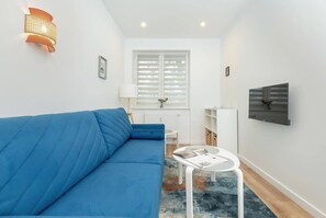 Un salon moderne avec un canapé bleu, une petite table blanche, un téléviseur fixé au mur et une fenêtre avec des stores blancs.