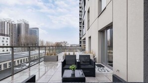 Un balcón moderno con mobiliario de mimbre negro, una planta en maceta y vistas a edificios de la ciudad.