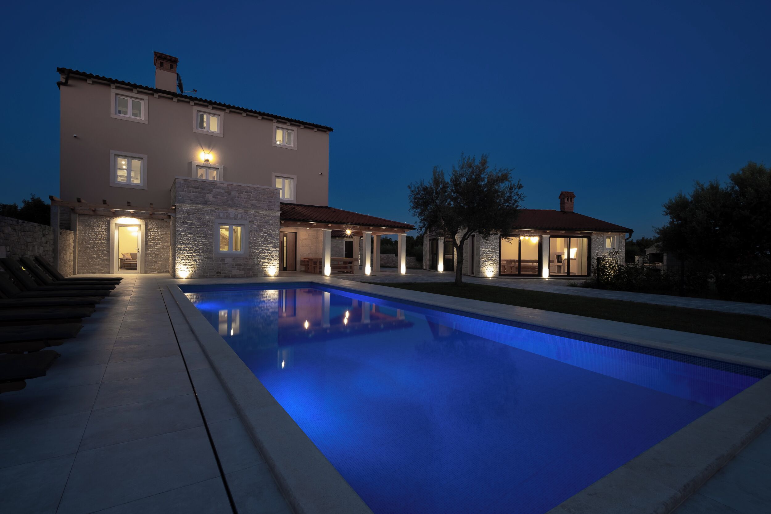 A modern house with a swimming pool, stone exterior, and multiple illuminated windows.