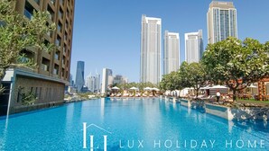A swimming pool with lounge chairs and umbrellas, surrounded by trees and buildings, including a tall EMAAR building.