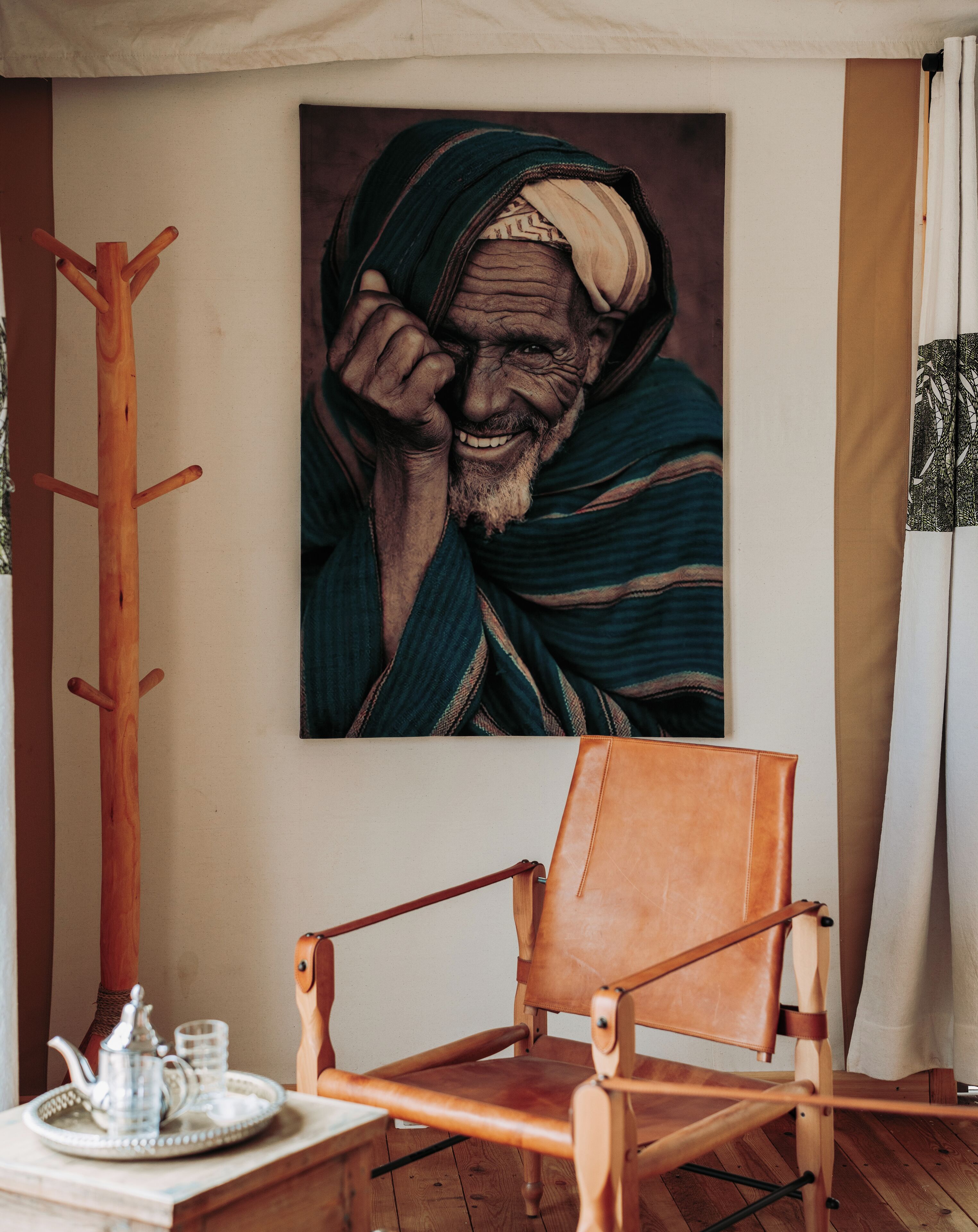 Una silla de madera con asiento de cuero, una bandeja metálica con tetera y vasos, y un cuadro de una persona en la pared.