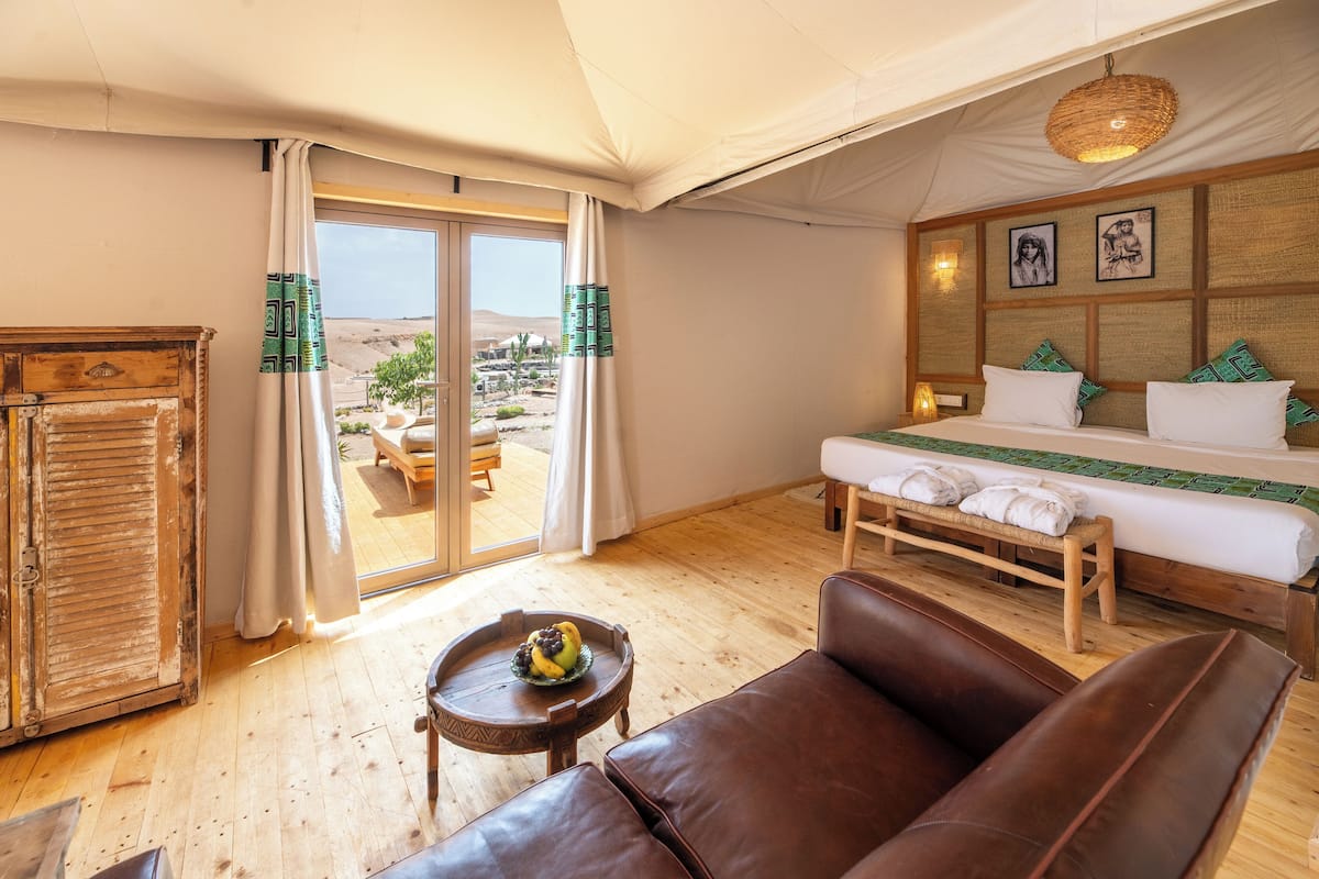 Una habitación con una cama, un armario de madera, un sofá de cuero, una mesita con frutero y vistas al desierto a través de una puerta corrediza.