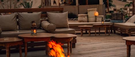 Un espacio al aire libre con un brasero, mobiliario de madera y faroles colgantes.