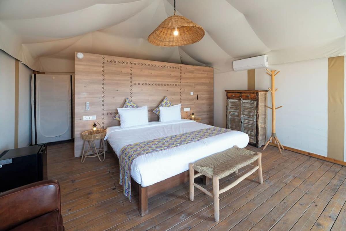 Un dormitorio con suelo de madera, una cama con edredón estampado, un banco tejido, una lámpara colgante y un armario.