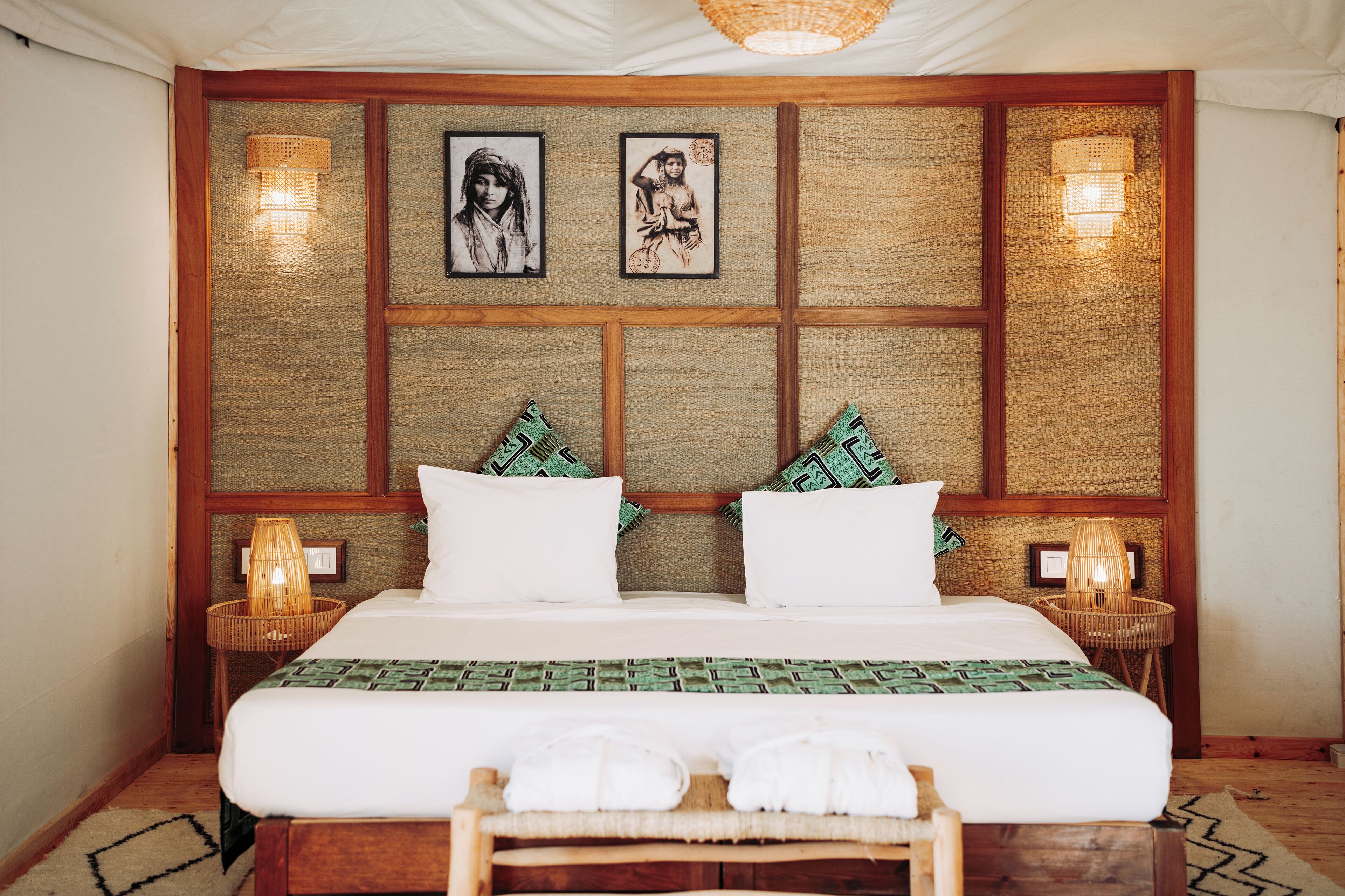 Una cama bien hecha con sábanas blancas, dos almohadas y una colcha verde estampada. Hay dos lámparas de pared a cada lado de la cama.