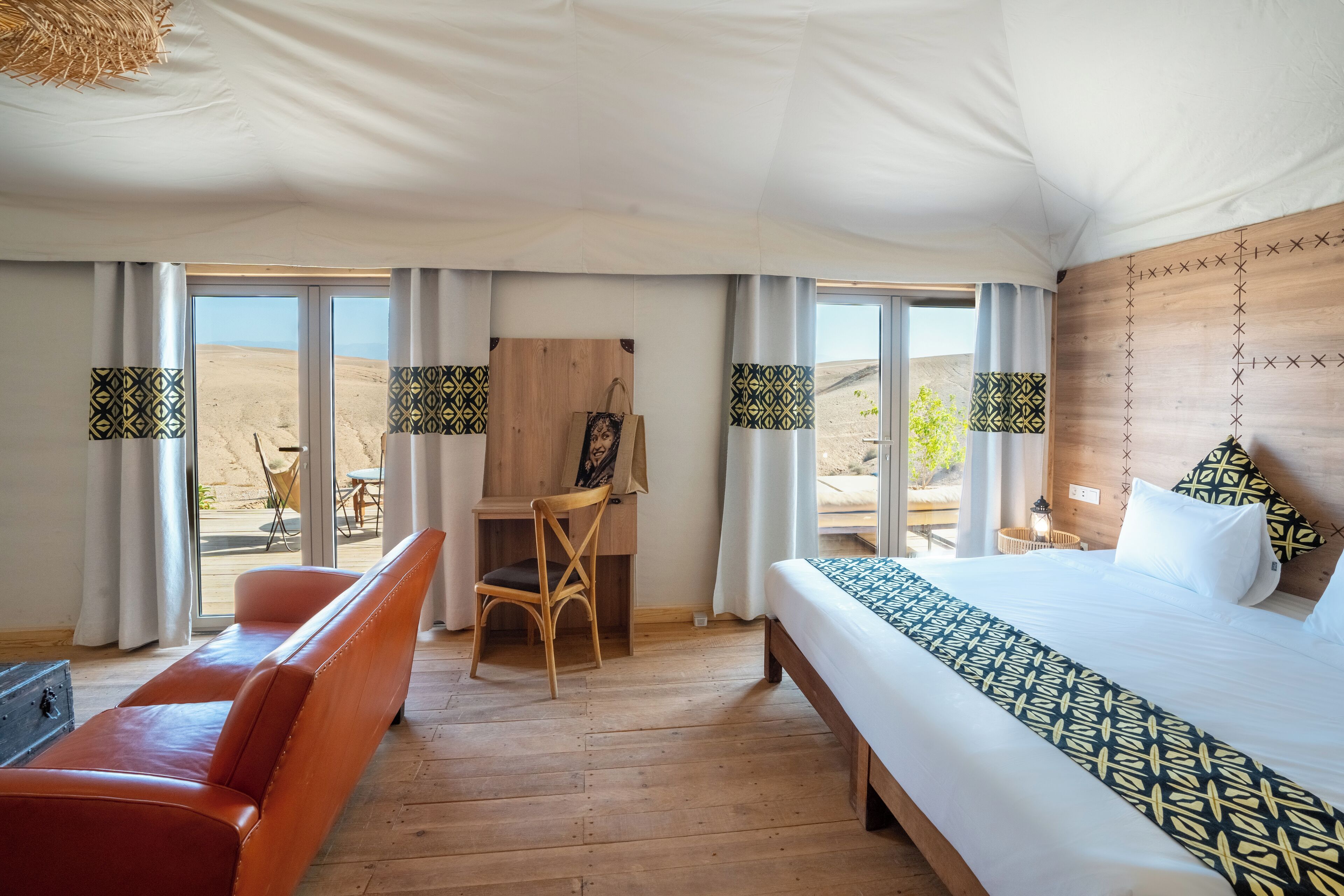 A room with a bed, a desk, and a chair. There are large windows with curtains.