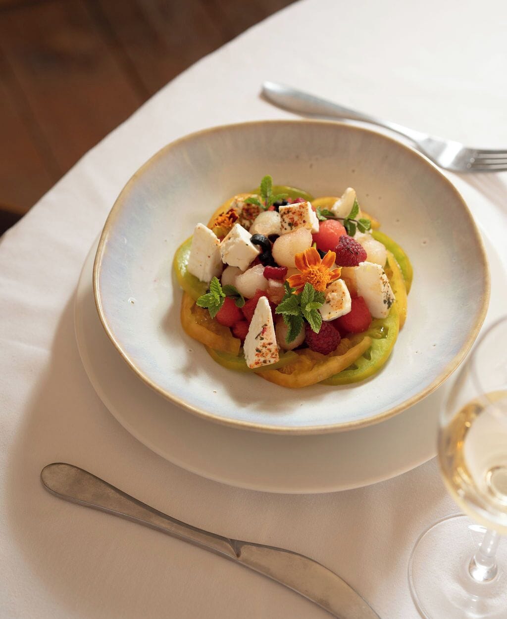 A plate of food with a variety of fruits and cheese on a white plate, accompanied by a fork and a glass of white wine.