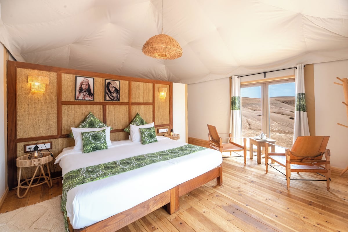 Un dormitorio amplio con una cama grande, cabecera de madera, panel decorativo tejido en la pared, mesita de mimbre, una silla de madera y vistas al desierto a través de la ventana.