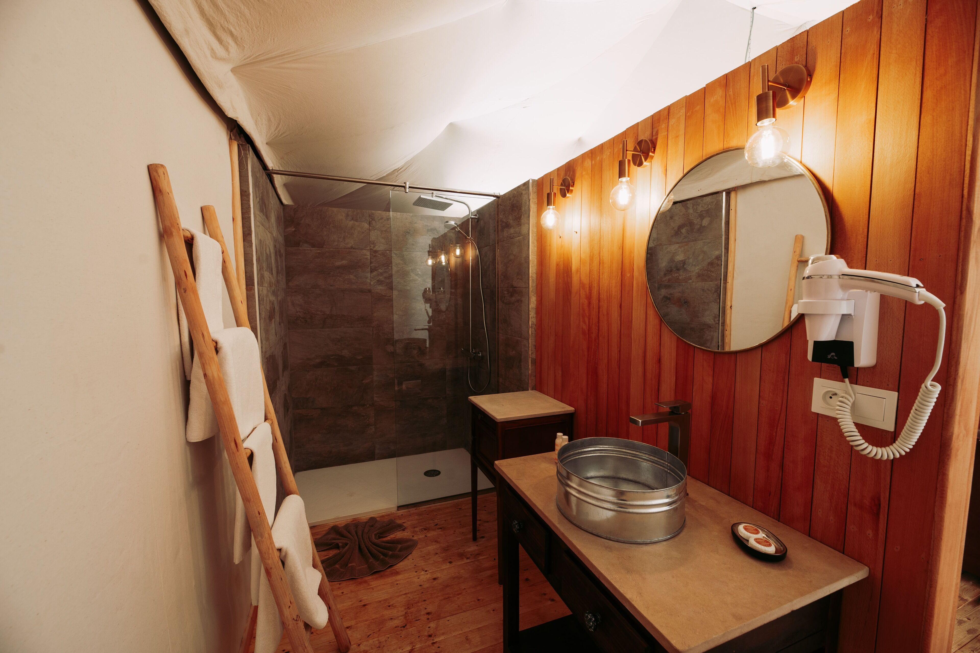A bathroom with a wooden wall, a round mirror, a hairdryer, and a metal basin on a wooden table.