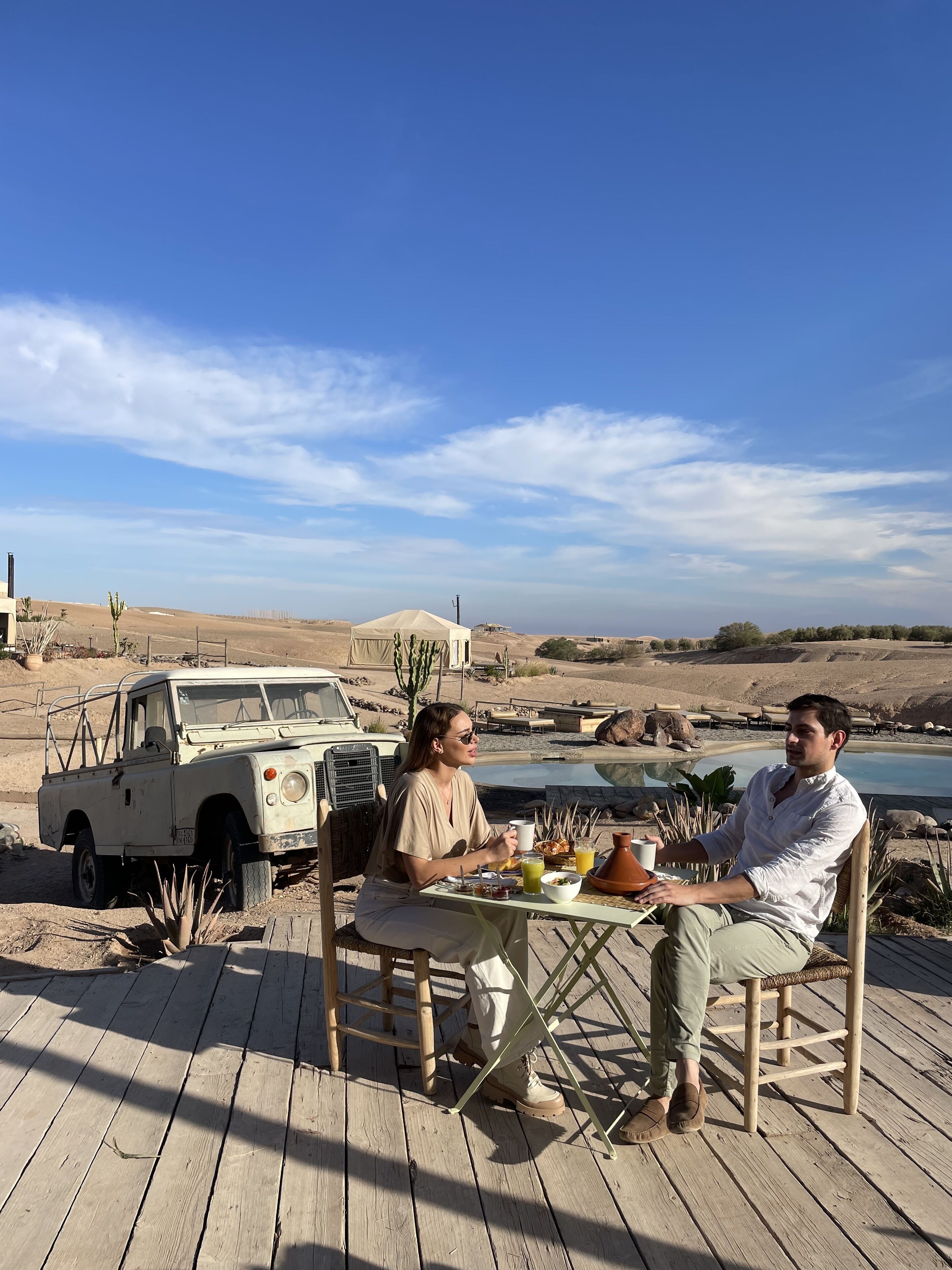 Two people dining at an outdoor table with a desert landscape and a vehicle in the background.