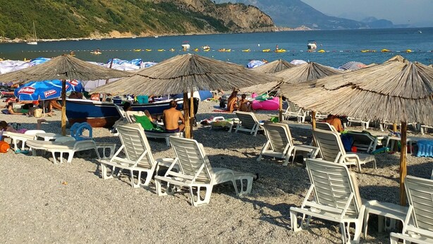 A beach with white lounge chairs and thatched umbrellas, a rocky hillside in the background, and calm sea. - Captivating and Spacious 2-bed House in Vranovici (Ljesevici)