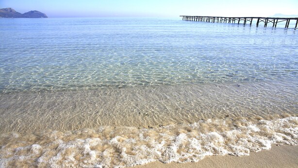 A beach with clear shallow waters, a sandy shore, and a pier extending into the sea.