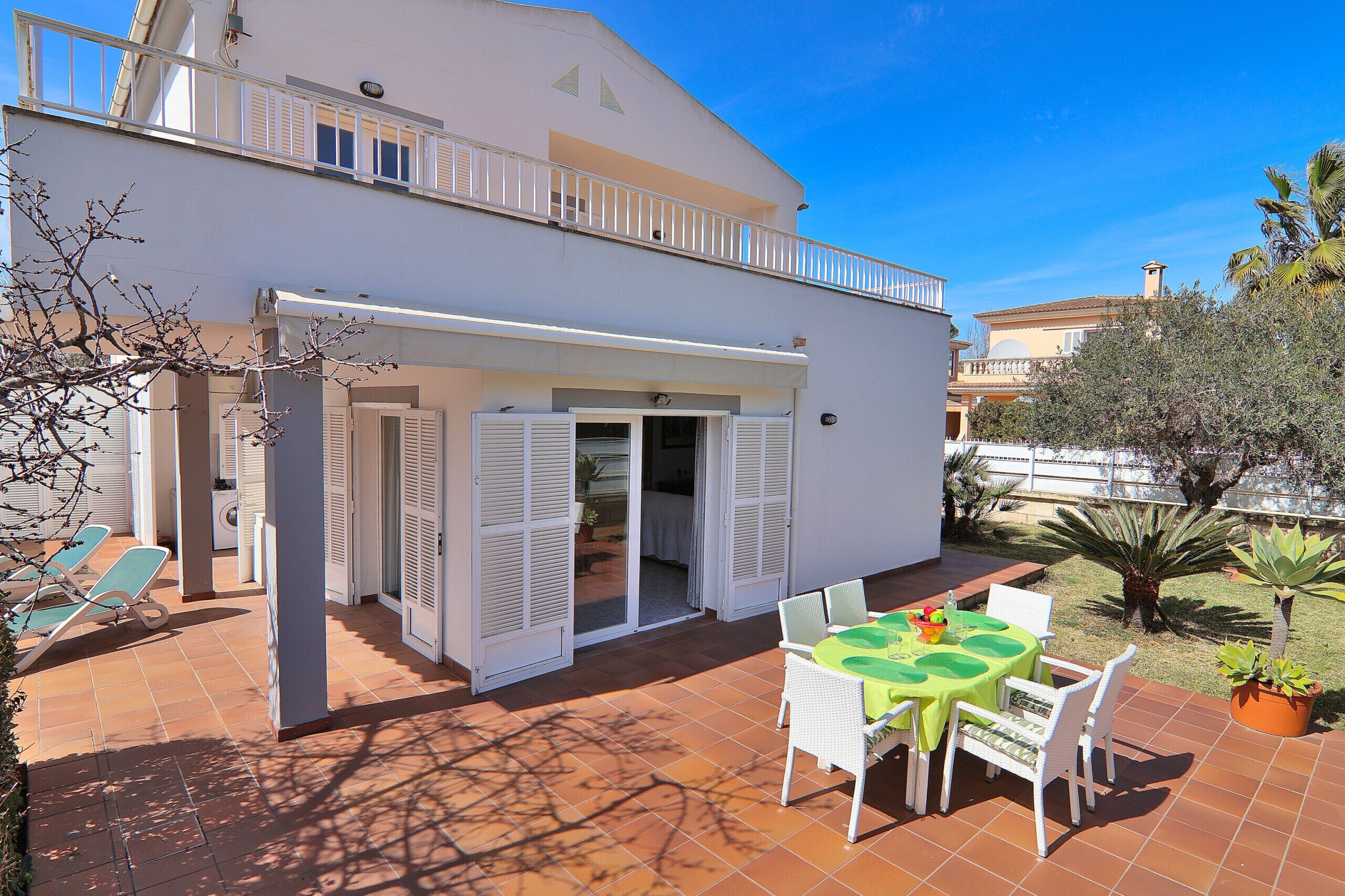 A two-story building with a balcony, a patio with a table and chairs, and a garden with palm trees.
