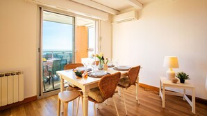 A dining area with a table set for four, wicker chairs, a small side table with a lamp, and a view through a glass door. - LE JARDIN BLEU AP4388 By Riviera Holiday Homes (Niza)