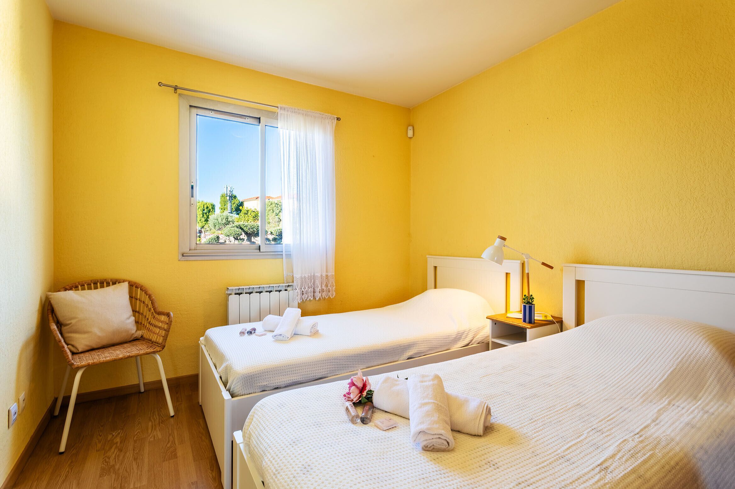 Une chambre d’hôtel avec deux lits, un fauteuil en osier, une fenêtre avec des rideaux et une petite table sur laquelle se trouvent une lampe et une plante.