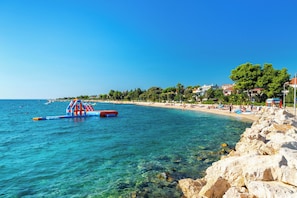 A coastal beach with clear water, a rocky shoreline, and a row of buildings in the background. - Room-private Room Near Center 6 (Novalja)