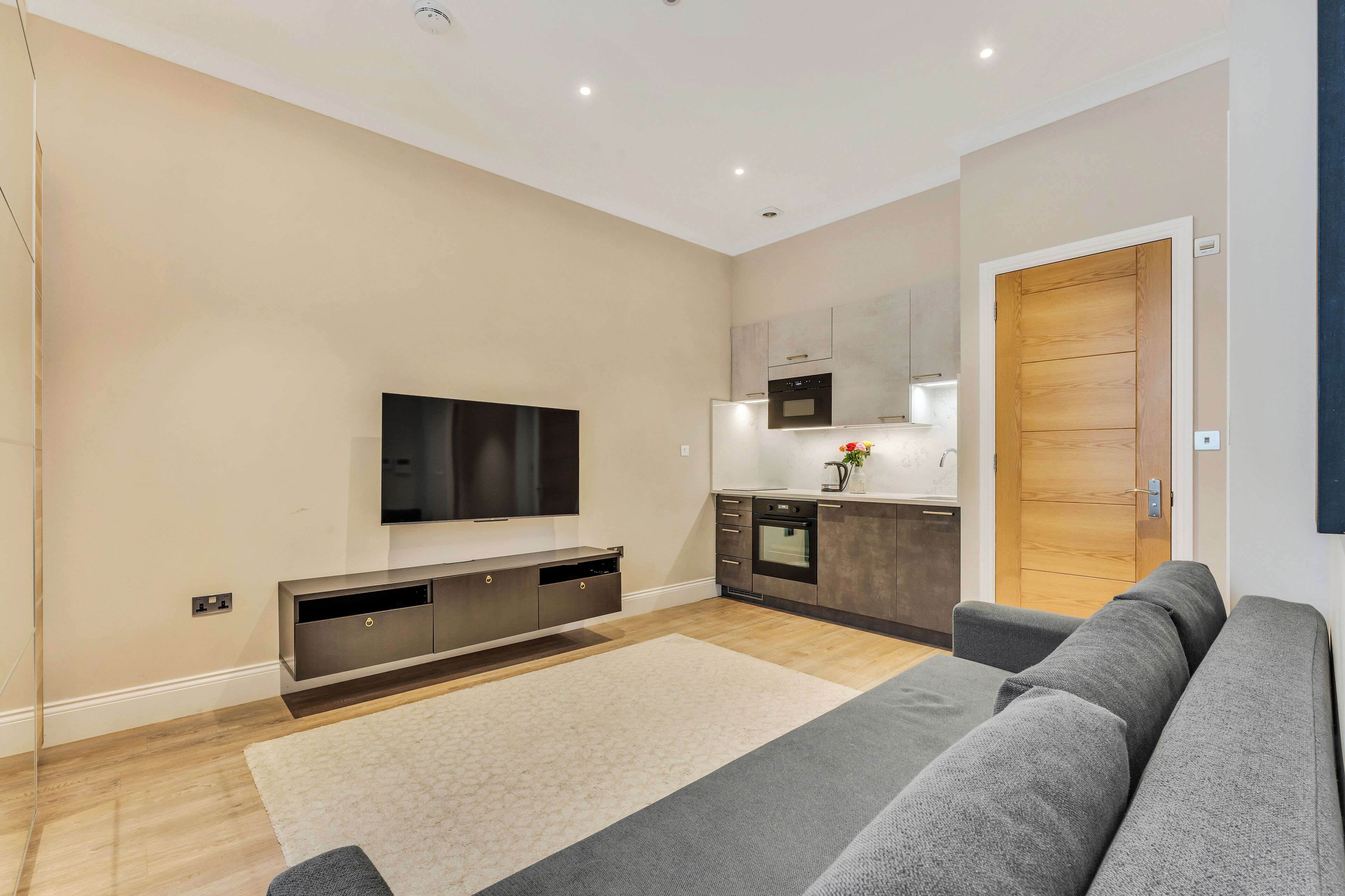 A modern living room with a flat-screen TV, grey sofa, wooden flooring, and a kitchen area with a microwave and oven.