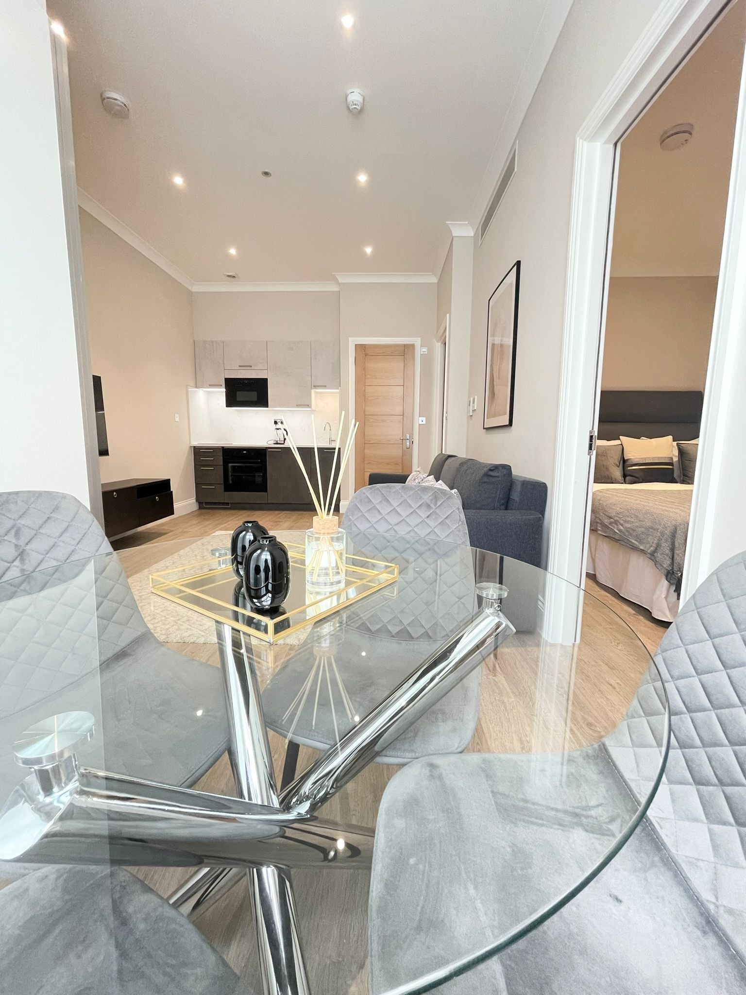 A modern living room with a glass coffee table, grey upholstered chairs, a sofa, and a kitchen in the background.