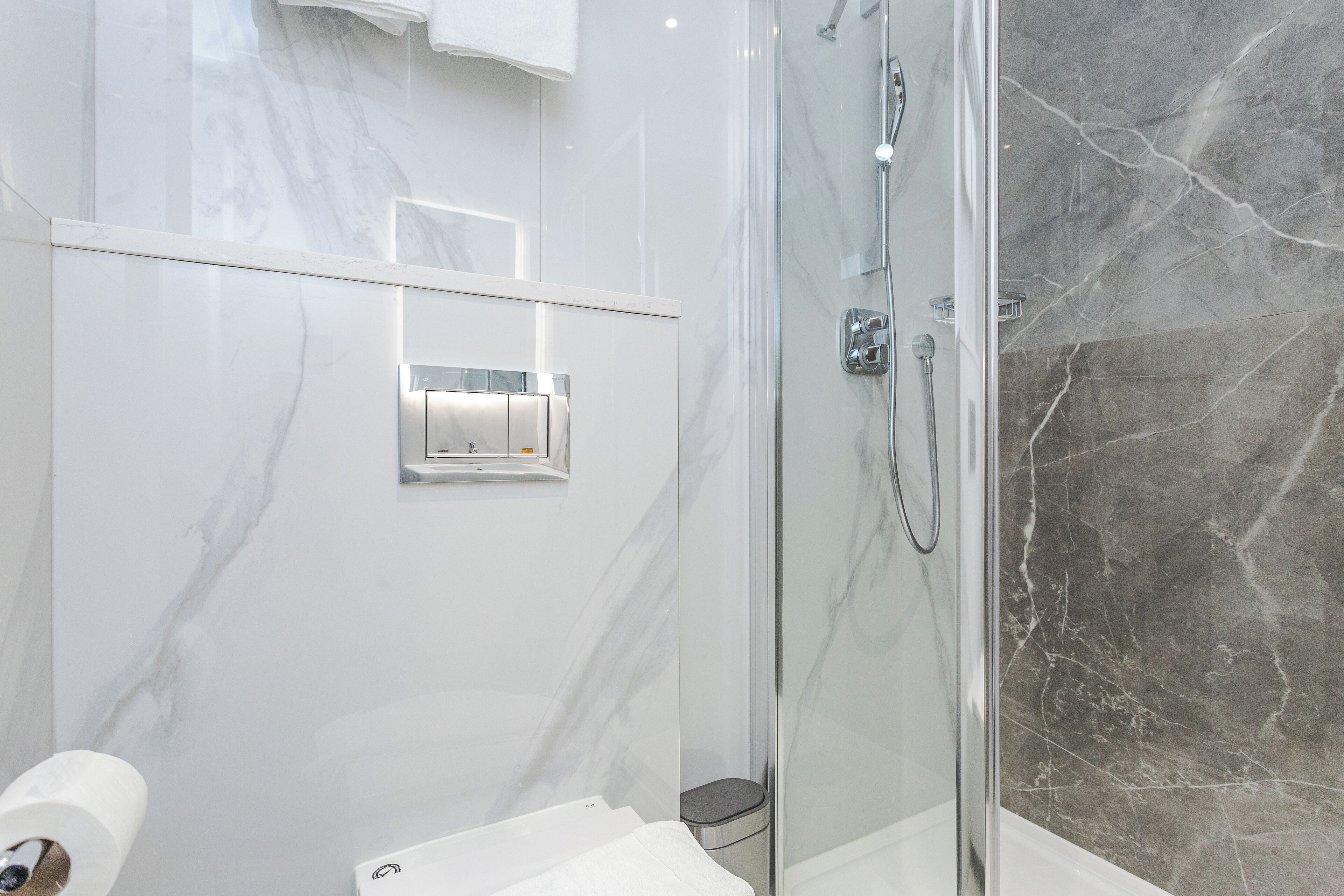 A modern bathroom with a glass-enclosed shower, marble walls, and a toilet with a paper towel dispenser.
