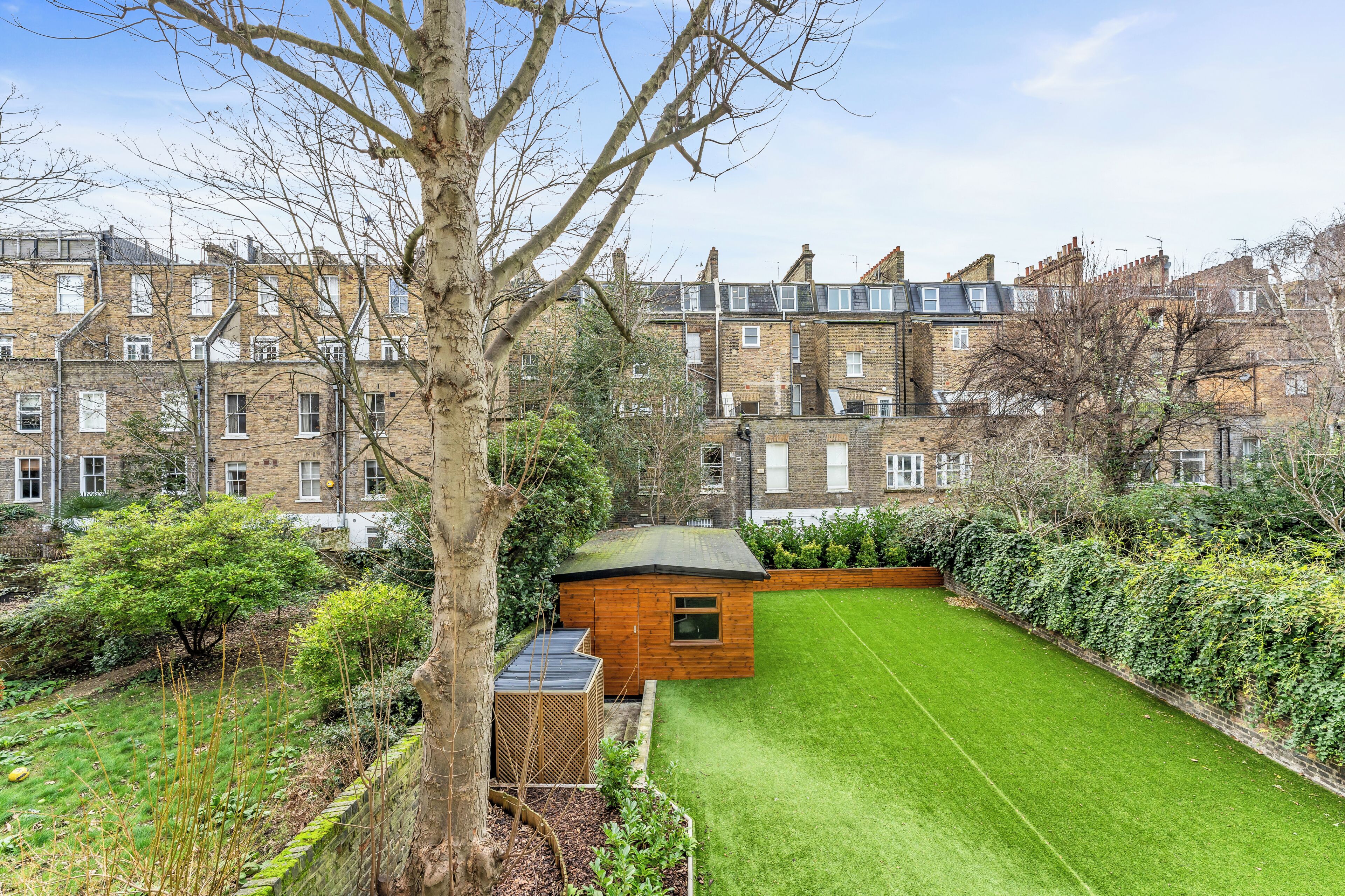 A residential area with a well-maintained lawn, a small wooden structure, and terraced houses in the background.