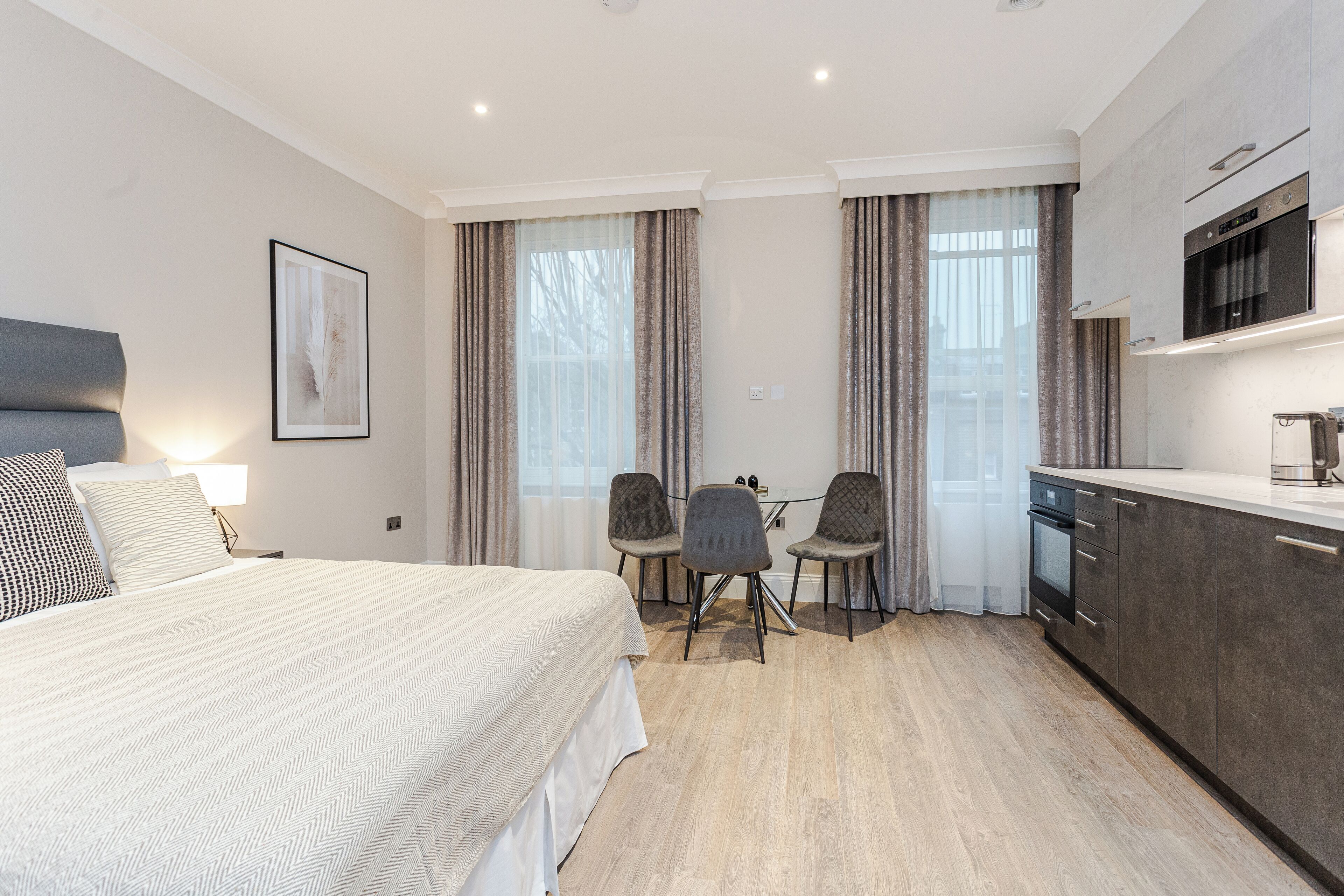 Ein modernes Hotelzimmer mit einem großen Bett, einem Schreibtisch mit zwei Stühlen, einer Küchenzeile und großen Fenstern mit Vorhängen.