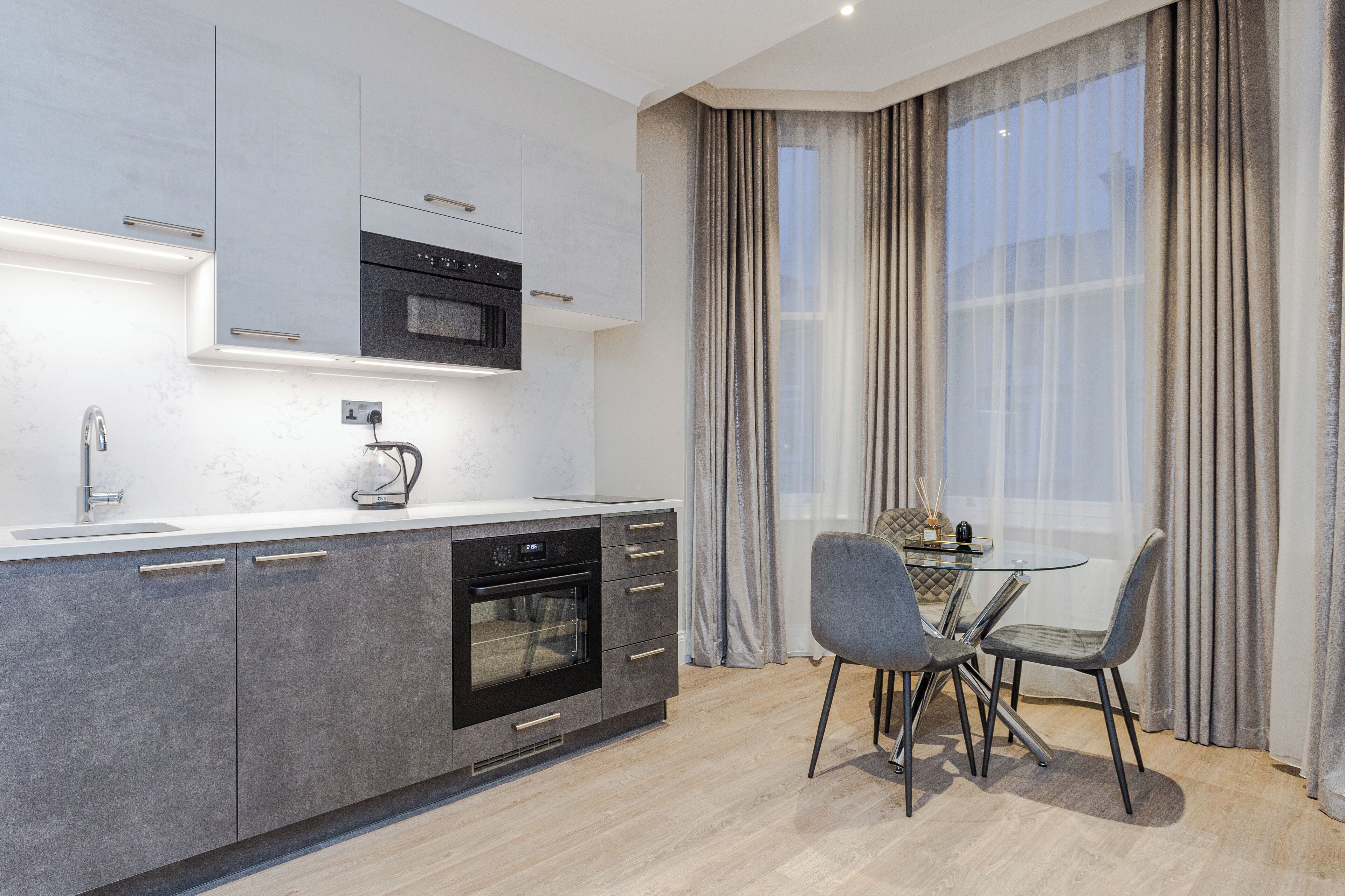 Una cocina moderna con horno empotrado, microondas y cafetera. Hay una mesa de comedor con sillas y una ventana con cortinas.