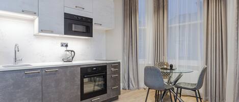 Una cocina moderna con horno empotrado, microondas y cafetera. Hay una mesa de comedor con sillas y una ventana con cortinas.